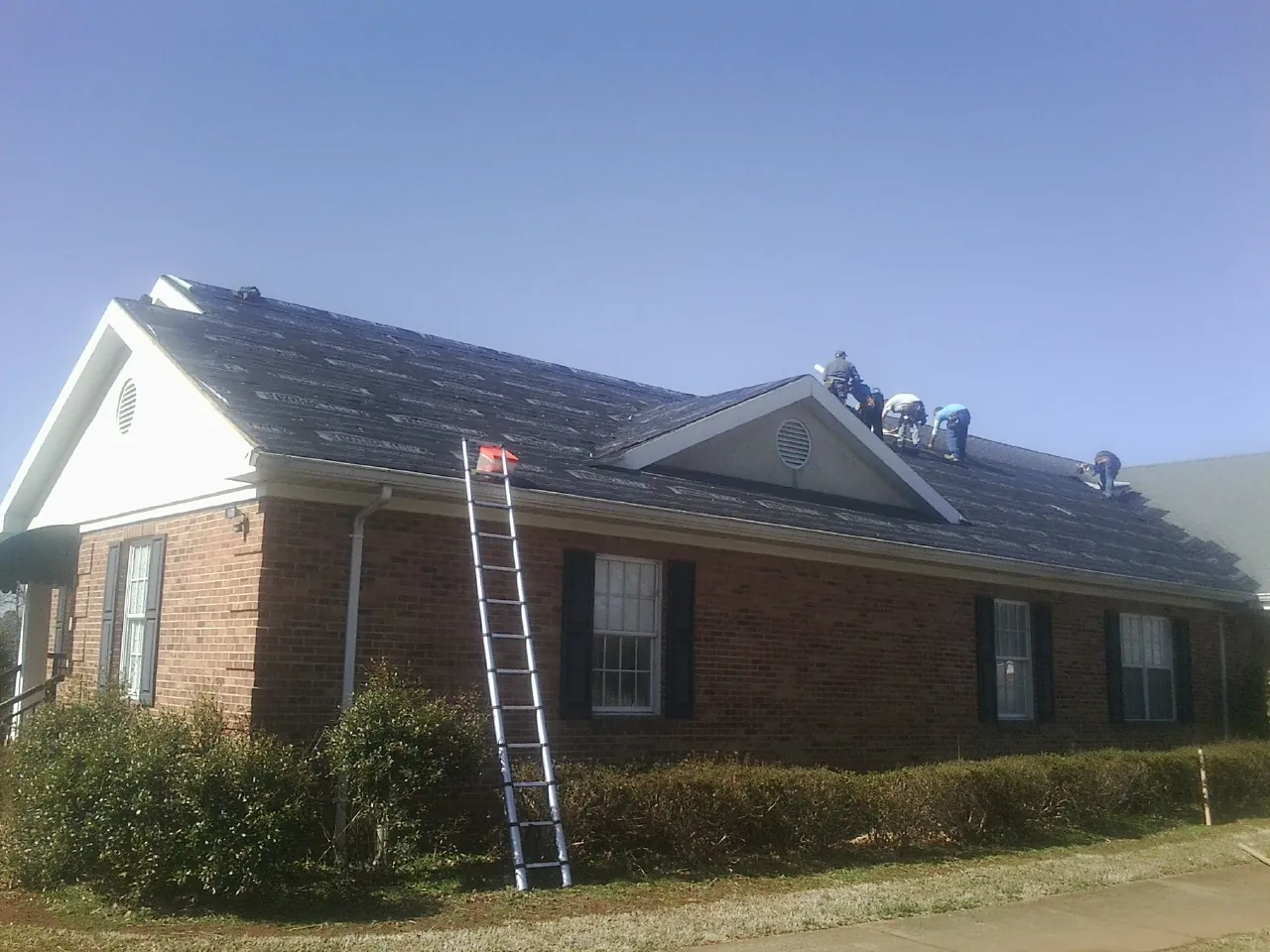 Crew tearing shingles off a brick ranch roof with a ladder set — Emergency Roof Repair work by our crew in Bleiblerville